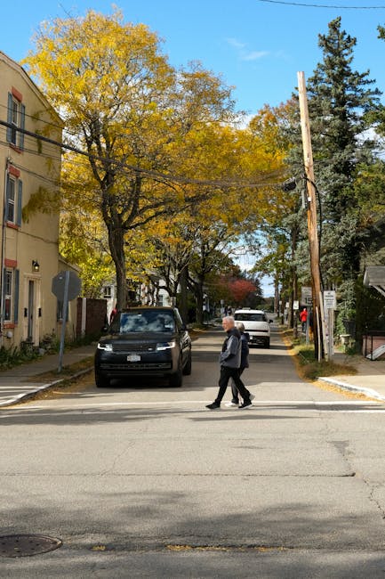 A street scene during daylight with a pedestrian crossing the road in the foreground, wearing a dark jacket and trousers. Behind him, there are parked cars including a black vehicle positioned close to the curb, and further along the street, other vehicles are visible parked on both sides. On the left, there is a yellow residential building with windows, and on the right, a mix of trees with autumn foliage and utility poles with overhead wires. The trees display yellow and green leaves, indicating the fall season. The sky is partly cloudy, and the street appears quiet, with no visible moving vehicles or pedestrians aside from the individual crossing the road. This setting could relate to a home relocation or moving process happening nearby, as part of local transportation and logistics services like those provided by Man with Van New Cross.