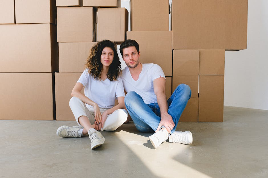 A young couple sitting on the floor inside a home surrounded by large cardboard boxes stacked against the wall, indicating an ongoing house move or packing process. The woman has curly brown hair and is dressed in a white T-shirt and light-colored pants, while the man has dark hair styled upward and is wearing a white T-shirt and blue jeans. Both appear to be taking a break during packing or furniture transport. Natural light illuminates the scene, casting shadows on the concrete floor. The boxes are sealed with packing tape and filled with household items for moving. This image depicts the practical aspects of home relocation, including the packing phase and the involvement of professional movers such as Man with Van New Cross, who handle furniture transport and packaging from inside the property to the vehicle, supporting efficient and secure moving logistics.