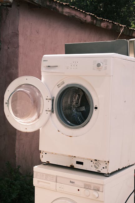 A white front-loading washing machine with its door open, positioned outdoors against a weathered pink wall under a sloped roof. The machine appears to be in the process of being prepared for removal, with no packaging or covering visible. The interior drum is visible through the open door, and the washing machine is placed on top of another appliance, possibly a dryer, indicating a home relocation or furniture transport scenario. The surrounding environment includes some greenery at the bottom left, and the image captures part of the loading process for a house move with the assistance of Man with Van New Cross, a professional removals service specializing in packing and moving logistics.
