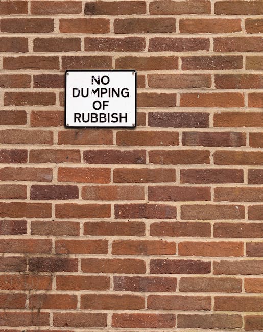 A rectangular white metal sign with black capital letters mounted on a red brick wall. The sign reads 'NO DUMPING OF RUBBISH' and is positioned approximately one-third from the top of the image. The bricks are uniform in size and colour, with light grey mortar between them, forming a neat, horizontal pattern. The sign is affixed directly to the brick wall, which appears clean and well-maintained. This image relates to house removals and moving logistics, emphasizing the importance of proper disposal and adhering to local disposal regulations as part of a professional relocation service. The scene indicates an outdoor setting, likely outside a residential or commercial property, and highlights the role of clear signage in managing rubbish disposal during home or business relocations.