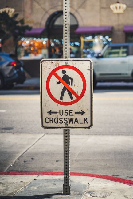 A rectangular traffic sign mounted on a metal pole situated on a city sidewalk in New Cross, indicating that pedestrians should not walk across the area, with the words 'USE CROSSWALK' and arrows pointing left and right. Behind the sign, a paved road is visible with parked cars on both sides, along with a building façade featuring large windows, purple awnings, and warm interior lighting. The sidewalk surface includes concrete slabs and a red-painted curb, and there are no pedestrians visible. The scene is captured in natural daylight, highlighting the urban environment typical of house removal and relocation settings, where clear signage assists in managing moving logistics and transport safety, as part of the local regulations and policies for safe furniture transport and home relocation.