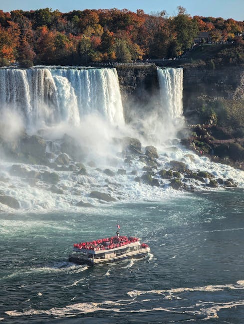 A boat carrying numerous red life jackets is moving across a river near a large waterfall. The waterfall is surrounded by dense trees with autumn foliage, and mist rises from the cascading water. The boat appears to be part of a sightseeing or transport service, navigating close to the rocky riverbank. This scene showcases a natural landscape with rushing water and lush greenery. In the context of house removals and furniture transport, a similar setting might involve the careful loading and transport of household items on boats or in vehicles navigating waterways, reflecting logistical aspects of moving services provided by Man with Van New Cross near Deptford Park to New Cross Gate area.