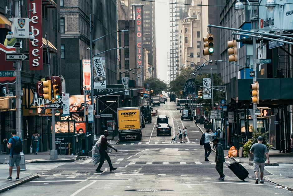 A busy city street scene in New York City with pedestrians crossing the intersection on a crosswalk. Several people are visible, some carrying backpacks or rolling suitcases, indicating a moving or relocation context. Vehicles, including a yellow Penske moving truck, are stopped at traffic lights near storefronts and high-rise office buildings. The scene includes various signs, traffic signals, and advertisements, with a prominent neon sign reading 'Radio City' visible above the street. The environment suggests a lively urban area with a mixture of commercial and pedestrian activity, where professional removals such as those provided by Man with Van New Cross may coordinate home or furniture transport through busy city streets.