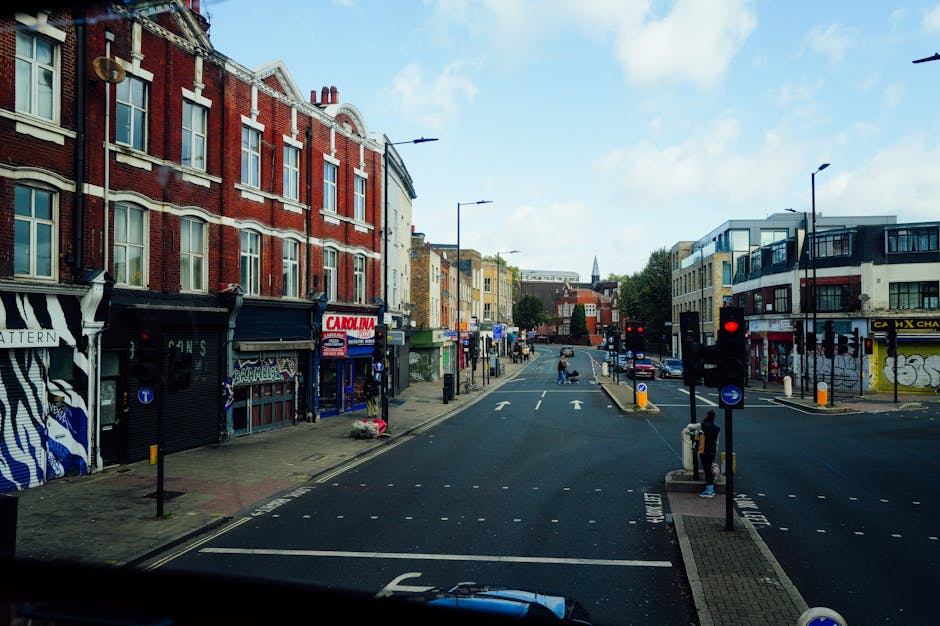 A daytime street scene in New Cross showing a busy intersection with multiple pedestrians walking along the pavement and crossing the road, which features white road markings and red dashed lines indi