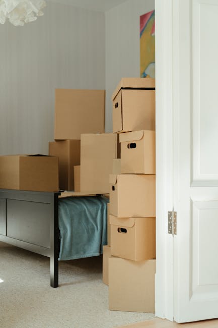 The image shows a collection of packed cardboard boxes of various sizes stacked on a dark wooden table with a light blue fabric covering part of the table's surface, inside a room with cream-colored walls and a decorative ceiling light. Some boxes are positioned directly on the table, while others are stacked vertically against a white door frame on the right side of the image, which is partially open. In the background, a colourful abstract artwork is visible on the wall. The scene appears to be part of a house removal or packing process, with [COMPANY_NAME] preparing items for transportation. The setting reflects typical interior space ready for a home relocation or furniture transport as part of a professional moving service offered by [COMPANY_NAME], aligned with the page titled 'SE14 basement flat removals: manoeuvring bulky items, NEW CROSS'.