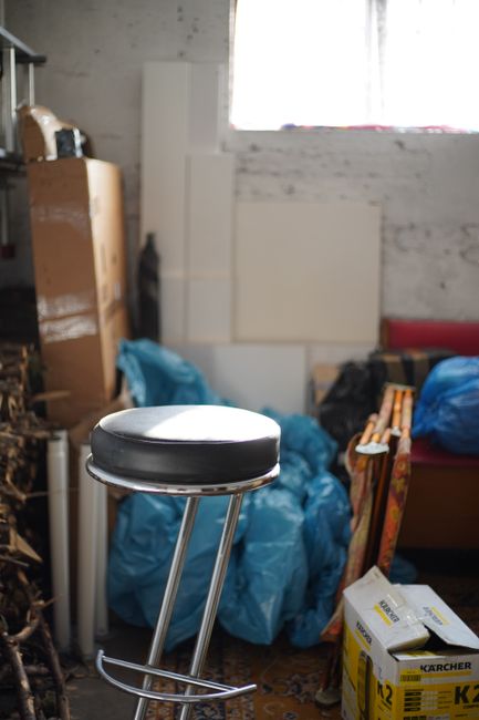 Inside a cluttered basement flat, a metal stool with a black cushioned seat is positioned in the foreground near a window with sheer white curtains, allowing natural light to illuminate the space. Behind the stool, there are multiple blue plastic bags filled with items, leaning against a white brick wall, along with stacked cardboard boxes and packaging materials. To the left, a wooden shelving unit holds various objects, while a cardboard box and a silver thermos or similar container are also visible. On the right side, a sofa or bed frame is partially seen, with a small white box of Kärcher cleaning equipment resting on the floor. The scene depicts a typical home relocation environment where packing and moving preparations are underway, and Man with Van New Cross’s removals services are assisting with the transportation of bulky items within the property.