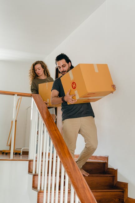Two movers from Man with Van New Cross are carrying cardboard boxes down a staircase inside a residential property during a home relocation. The man in the foreground, wearing a dark t-shirt and beige trousers, holds a medium-sized cardboard box secured with tape in front of him as he descends wooden stairs. Behind him, a woman with curly hair, dressed in a light-colored top, carries another cardboard box. The staircase features wooden steps with a white wall to the side, and a wooden handrail attached to posts. The interior space is well lit with natural light, and the environment indicates a careful packing and safe handling process as part of furniture transport and moving logistics for a house removal. This scene exemplifies the loading process within a moving service provided by Man with Van New Cross, focusing on efficient and secure handling of household items during a domestic move.