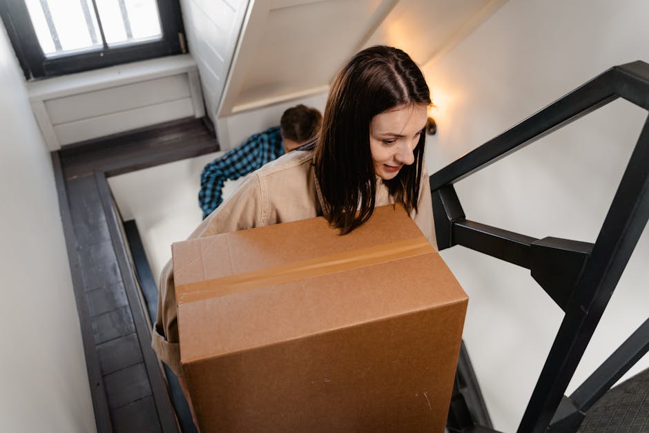A young woman with shoulder-length brown hair, dressed in a beige top, is carrying a large cardboard box up a narrow staircase within a residential property during a home relocation. Behind her, a man wearing a blue checkered shirt is also ascending the stairs, likely assisting with furniture transport and packing. The staircase has dark wooden steps, a black handrail, and white walls, with natural light coming through a window at the top. The environment suggests an interior space prepared for a professional move, and the visible items include the cardboard box, padding materials, and the move equipment such as the handrails. This scene reflects the process of packing and loading during a house removal, with the team from Man with Van New Cross working to safely navigate the tight staircase in a typical London home.
