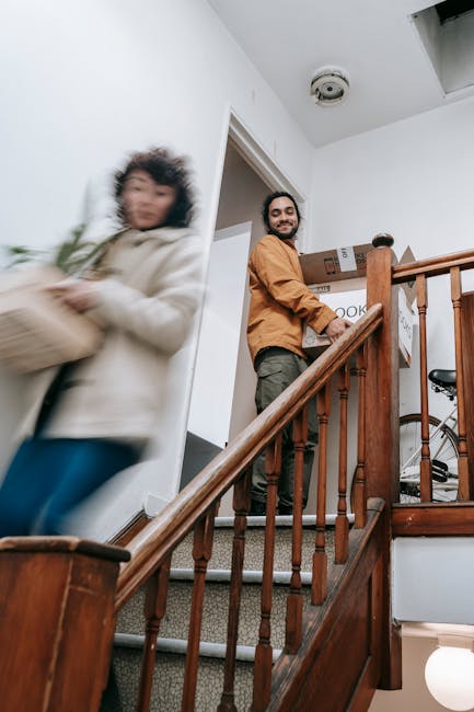 Two movers from Man with Van New Cross are carrying cardboard boxes down a staircase inside a residential property during a home relocation. The man in the foreground, wearing a dark t-shirt and beige trousers, holds a medium-sized cardboard box secured with tape in front of him as he descends wooden stairs. Behind him, a woman with curly hair, dressed in a light-colored top, carries another cardboard box. The staircase features wooden steps with a white wall to the side, and a wooden handrail attached to posts. The interior space is well lit with natural light, and the environment indicates a careful packing and safe handling process as part of furniture transport and moving logistics for a house removal. This scene exemplifies the loading process within a moving service provided by Man with Van New Cross, focusing on efficient and secure handling of household items during a domestic move.