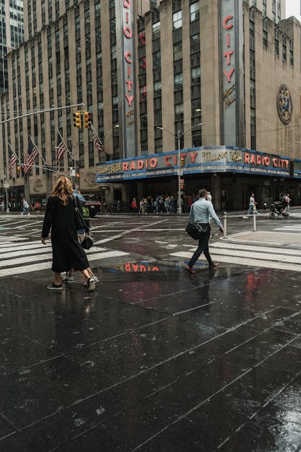 A street scene in front of Radio City Music Hall in New York City on a rainy day. The theatre's marquee displays the venue name in bright blue and yellow lights, with the classic neon sign reading 'RADIO CITY' vertically on the building's facade. Two pedestrians, a woman with long brown hair dressed in dark clothing and a man with short hair wearing a grey jacket and black pants, walk across the wet, reflective pavement holding bags. The street is lined with traffic lights, flags, and other urban elements. The surrounding buildings are tall, made of concrete and glass, indicative of a busy downtown area. The scene captures a moment of home relocation or moving logistics outdoors, with the emphasis on the urban environment and pedestrian activity.
