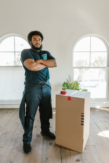 A man with a dark afro hairstyle and beard, dressed in dark clothing, stands indoors with arms crossed in front of a large cardboard box sealed with red packing tape. The box is positioned on a wooden floor near a white wall with two large, arched windows allowing natural daylight to fill the room. Outside the windows, trees and parked cars are visible. A green potted plant with broad leaves is placed behind the box, and some packing tape and cardboard pieces are visible nearby on a surface. The scene suggests an ongoing home relocation or packing process, reflecting the loading or packing phase of house removals. This image is associated with Man with Van New Cross, a company specializing in local removals, emphasizing professionalism and careful handling of belongings during moving services.